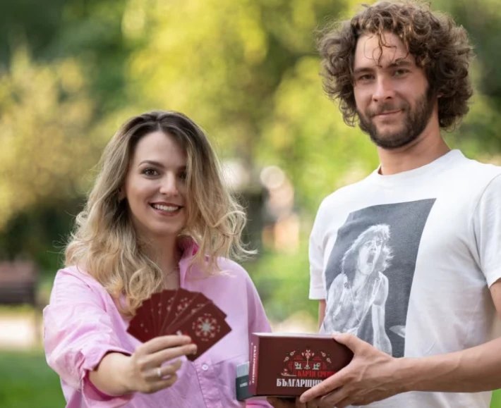 Two people holding Българщината cards and the game box outdoors